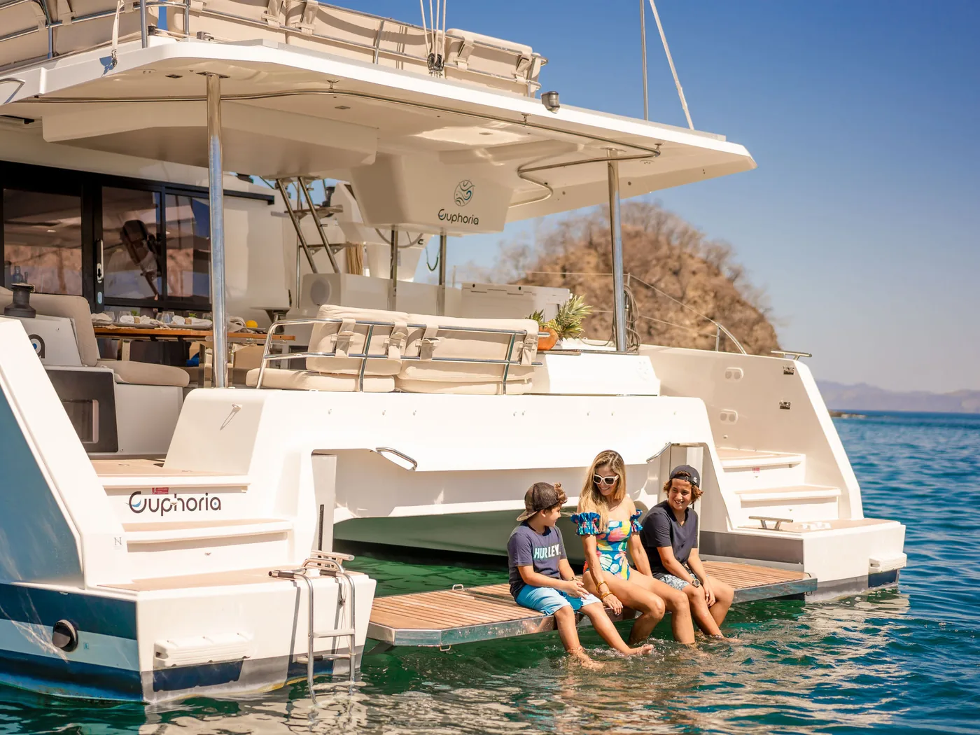 Guests sitting on the transom steps of an Ocean Club catamaran in clear turquoise water, Costa Rica